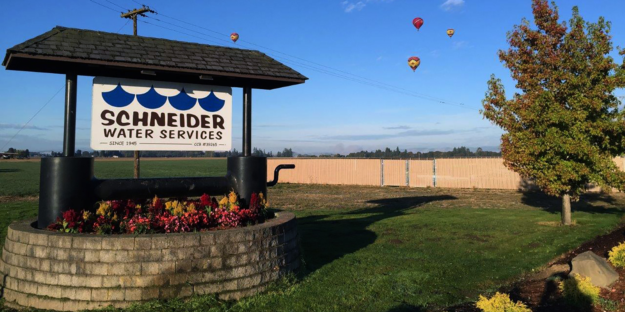 Schneider Water Services sign in a green patch of grass with the blue sky behind and four hot air balloons in the air